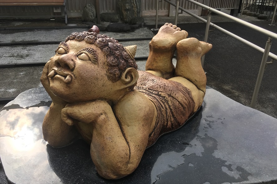 くろがね橋近くにある足湯「鬼怒子の湯」の前で寝そべる遊心鬼。片方のツノにリボンを結んだ、おしゃれな女の子の鬼 